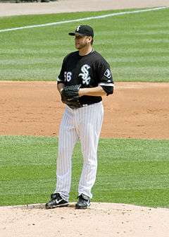 A man standing on a pitcher's mound holds a baseball in his black baseball glove. He is wearing a black-and-white baseball uniform.