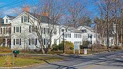 A row of two-story white houses with black or gray gabled roofs down a street on the left of the image. In the rear, slightly obscured by some bare trees, is a similar church with a short four-pointed steeple.