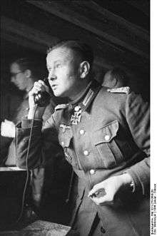 Black-and-white portrait of a man on the phone in semi profile wearing a military uniform with an Iron Cross displayed at his neck. He is holding a cigarette in his left hand.