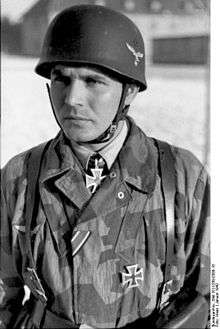 A man wearing a camouflage military uniform, steel helmet and a neck order in the shape of a cross.