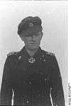 A man wearing a military uniform, peaked cap and a neck order in the shape of a cross. His cap has an emblem in shape of a human skull and crossed bones.