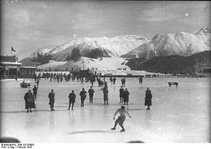 A male figure skater performing on a large frozen outdoor area with spectators and judges nearby on the ice. The background shows snow covered mountains and a building.