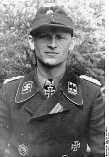 A black-and-white photograph of a man wearing a military uniform, cap and a neck order in shape of an Iron Cross. His cap has an emblem in shape of a human skull and crossed bones.