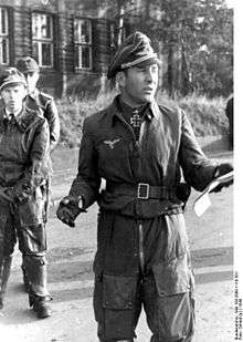 Black-and-white portrait of a man wearing a peaked cap and military flight suit with an Iron Cross displayed at his neck. He is holding a sheet of paper in his left hand and appears to be talking to a larger audience.