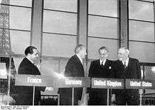 Four men stand behind podiums with their country names of France, Germany, United Kingdom and United States, in front of a backdrop of the Eiffle Tower.