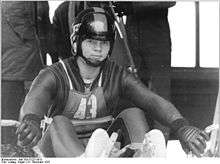 A man wearing a skin-tight cold-protecting jumpsuit, gloves, and a full-face helmet with a lifted visor. He is sitted on a sled and there are people behind him.