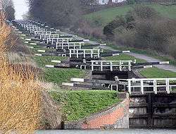 A series of approximately 20 black lock gates with white ends to the paddle arms and wooden railings, each slightly higher than the one below. On the right is a path and on both side's grass and vegetation.