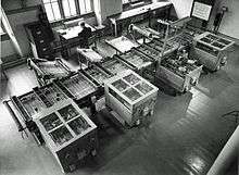 Looking down from above at a room at an intricate mechanical device which fills the room. In the background, a man sits at a desk next to a filing caninet.