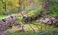 Two intersecting stone walls below a larger stone retaining wall in a grassy, shrubby area. There is a small set of table and chairs next to a large vase in a cleared brick area at the corner