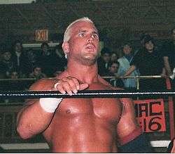 A white male standing in a wrestling ring with black ropes with his mouth open wearing wrestling gear in front of a crowd.