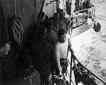 A bearded man wearing a white jumper walking towards the camera along a narrow walkway while holding on a railing. Water is visible below the walkway and a large number of men wearing dark clothes are either passing in the opposite way or watching the bearded man.