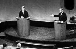 Two men stand at podiums on a stage. The man on the right is speaking while gesturing to the man on the left. Two other men are seated, facing the podiums.