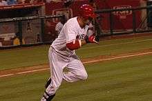 A dark-skinned man with "NGEL" visible in red text on a white jersey runs from home plate to first base. He has on red and black batting gloves, a red batting helmet, white pants and black shinguards.