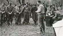 A chaplain reads from a bible while a formation of men stand in the background with heads bowed.