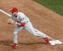 A left-handed man in a gray baseball uniform with red trim and a red baseball cap stretches to reach a ball with his tan baseball glove; his foot is resting on a white square base.