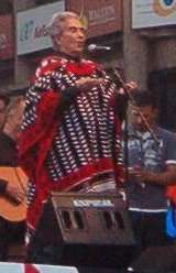 A woman standing behind a microphone on a stage; she is wearing  colorful outfit and has her arms bent so that her hands are at chest-level.