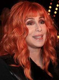 A Caucasian female with shoulder-length hair chemically dyed auburn with cool, brown colored makeup smiles, looking off to the right in front of a black background.