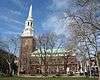 Ground-level side view of a brown brick church with a large, white, tapering spire.