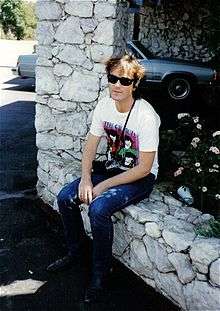 Steve Kilbey is sitting on a low rock wall beside a garden bed and a rock pillar. He is wearing dark glasses, a white tee-shirt with The Church and four faces (partly obscured), and jeans.