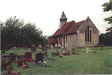 A small stone church with red tiled roofs seen through a churchyard from the southeast. On its far gable is a bellcote with a pyramidal roof