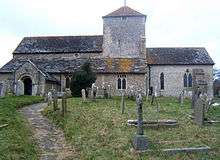 A long church with flint walls and tiled roofs. A short tower with a pyramidal roof and two small windows rises slightly right of centre. To the left, a longer section with a high roof, in front of which is a projection with a lower roofline and two low windows. A porch with an arched doorway extends from the far left side. To the right, there is a shorter and lower section with two windows. A path leads through a graveyard to the porch.