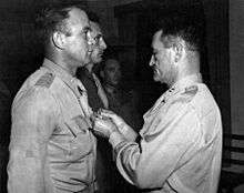 A monochrome photograph of several military men standing at attention, with one lit prominently in the foreground on the left, and an older man prominent on the right wearing the uniform of a two-star general, pinning a medal on the first man's uniform