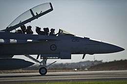  Forward section of military jet on an airfield, with open canopy and two crewmen seated in the cockpit