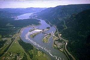 Aerial photograph of the Bonneville Dam complex on a sunny day, including the dam itself, facilities and highways on both sides of the Columbia River, and the rugged scenery of the Columbia Gorge.