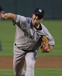 A man in a gray baseball uniform with "New York" across the chest in navy-blue letters and wearing a navy blue baseball cap with an interlocking white "NY" on the front