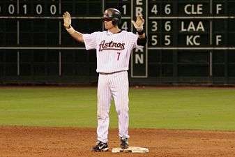 A man in a white, pinstriped baseball uniform with "ASTROS 7" on the chest and a black batting helmet stands on a base with both hands raised.