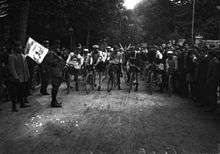 A group of cyclists standing behind a person with a flag, with other people standing next to the road