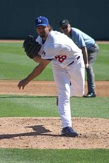 A man wearing a white baseball uniform and a blue baseball cap follows through after throwing a baseball