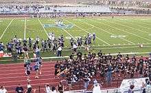 A high school football field with a large "DV" emblem in the center of the turf. Dougherty Valley football players in blue and silver uniforms are lined up on the side of the field. On the track surrounding the field, there are cheerleaders in light blue, white, and navy blue uniforms with blue pom-poms to the left. To the right of the track there are members of the pep band with their instruments in black shirts.