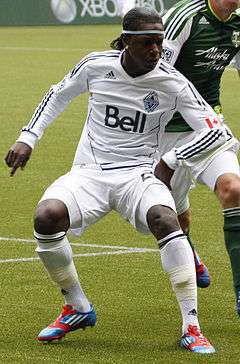 A man wearing a white long sleeve shirt, white shorts, white socks, and red and blue shoes, in motion on a soccer field, looking to his left. In the background, a man wearing a green shirt, white shorts, and green socks.