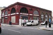 A red tiled building sited on a corner of a road junction. Five large, semi-circular windows fill much of the upper storey with the two on the corner removed and replaced with ventilation grilles.