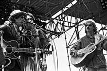 On stage outdoors, Dylan in a leather jacket and iconic porkpie hat & scarf, Baez wears a peasant skirt, sarape, tassled scarf around neck, her arm around Dylan's shoulder. Santana in workman's shirt & knit cap.