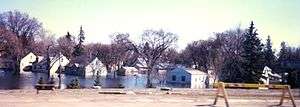 Several homes surrounded by water. Large trees are located nearby. A safety construction barrier can be seen at the right of the image.