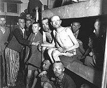 A group of malnutrished prisoners, some of them naked, some dressed in prisoner clothing, fill the interior of one of the barracks, as well as two lower levels of a three-storey bed.