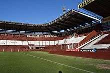 Corner of a stadium with empty maroon and white seating sections, viewed from the pitch, from which it is separated by a fence; the sky is blue and cloudless.