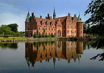 A large three-storied castle has  steeply pitched roofs,  two circular towers and a spire, and a large arched entrance facing a lake which reflects the castle's image.