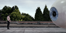 A man stands on a rooftop across from a giant eyeball.