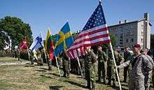 Six flags are held by a line of soldiers in camouflage as they stand in front of trees and a beige building.