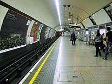 A wide concrete platform in a circular tunnel. Railway track runs along the left with posters fixed to the wall opposite the platform.