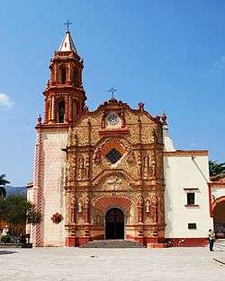 Facade of a church with one tower. The main portal is richly decorated with relief and sculptures and features a clock at the top.