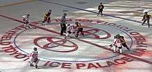 In the middle of a large red circle with the Toyota logo, lit partially by sunlight shining through the roof above, two hockey players from different teams are attempting to take control of the puck beneath them. The referee, in a black and white vertically striped shirt with an orange band around his upper right arm, stands off to their left. Around the edges of the circle are the two players' teammates, some just beginning to skate into the circle.