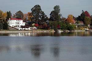  Houses with neat lawns, gardens, and, in some cases, boat docks, face a lake. A white, three-story house with a red roof stands out because of its size and color and because the other houses are mostly hidden by trees and shrubbery. Some of the foliage is red or golden, the rest green. The lake is calm, the sky clear.