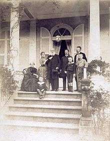 Photograph of a group of people assembled on a columned porch at the top of a flight of steps, with one older lady seated, one younger lady leaning on the arm of an older bearded man, two younger men and three small boys