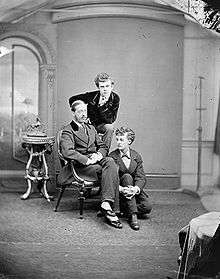 A studio, black and white photograph of three men, one is seated on an armchair, another is sitting on the floor beside him. The third is sitting behind them on a high stool and looks into the camera.