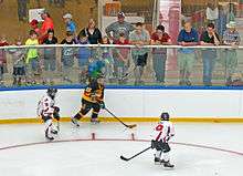 A hockey player in a black uniform with a puck on his stuck is confronted by two opponents in white near the wall of the rink. Above him, adults and children variously dressed in warm-weather clothing such as T-shirts, shorts and sandals watch them closely from between the glass and netting behind it