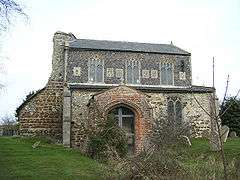 A stone church with a brick porch seen from the south. To the left are the remains of the collapsed tower. The clerestory contains three windows and six carved panels.